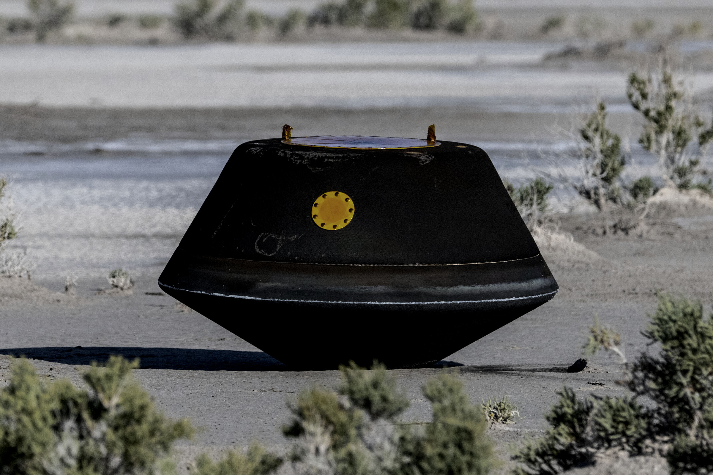 A small reentry capsule rests on desert ground after landing, its ablative heat shield charred from atmospheric entry.