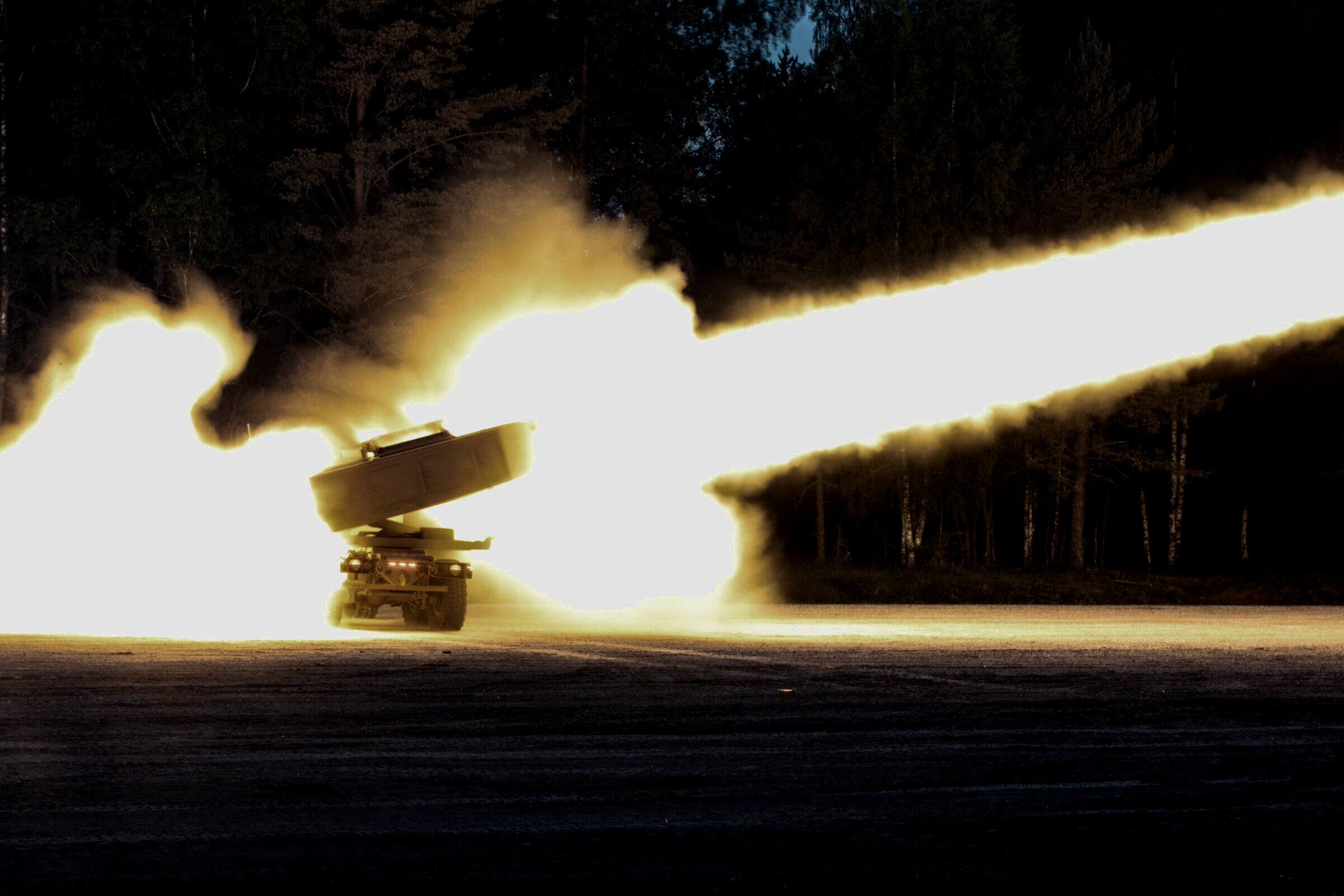 A launcher fires a rocket at night, the motor plume streaking across a dark treeline.