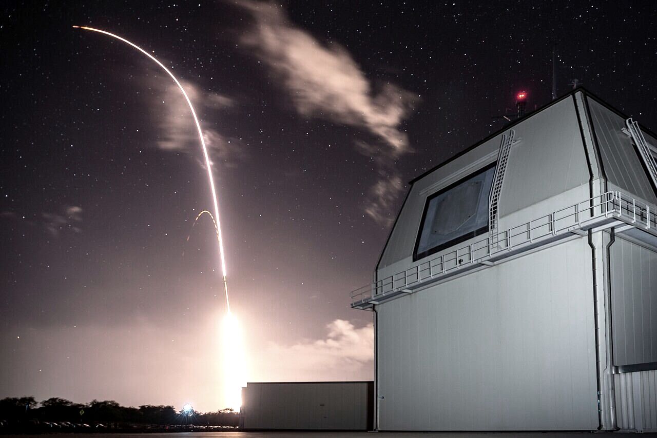 A long-exposure night photograph of an interceptor missile arcing into a starry sky above the Aegis Ashore test complex.